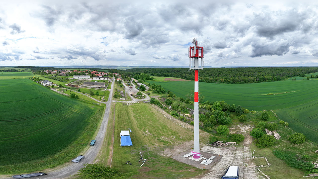 Weitwinkelaufnahme eines hohen rot-weißen Mobilfunkmasts und einer künstlichen Baumspitze zur Tarnung. Im Hintergrund sind Felder, Wälder und ein kleines Dorf zu sehen.