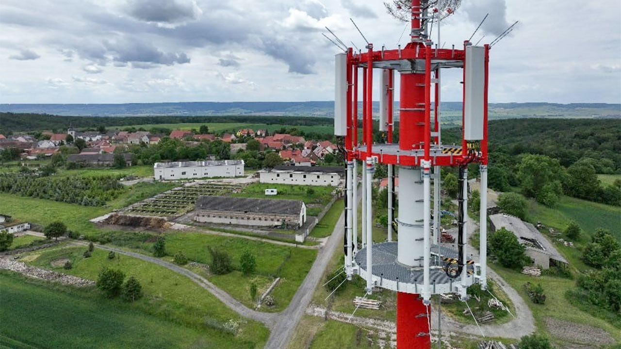 Nahaufnahme eines rot-weißen Mobilfunkmasts. Im Hintergrund sind Felder, Wälder und ein kleines Dorf zu sehen.