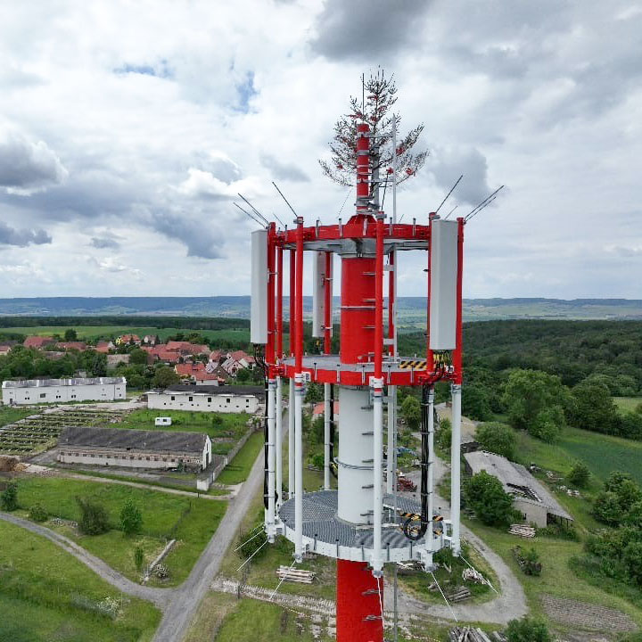 Nahaufnahme eines rot-weiß gestrichenen Mobilfunkmasts und einer künstlichen Baumspitze zur Tarnung. Im Hintergrund sind Felder, Wälder und ein kleines Dorf zu sehen.
